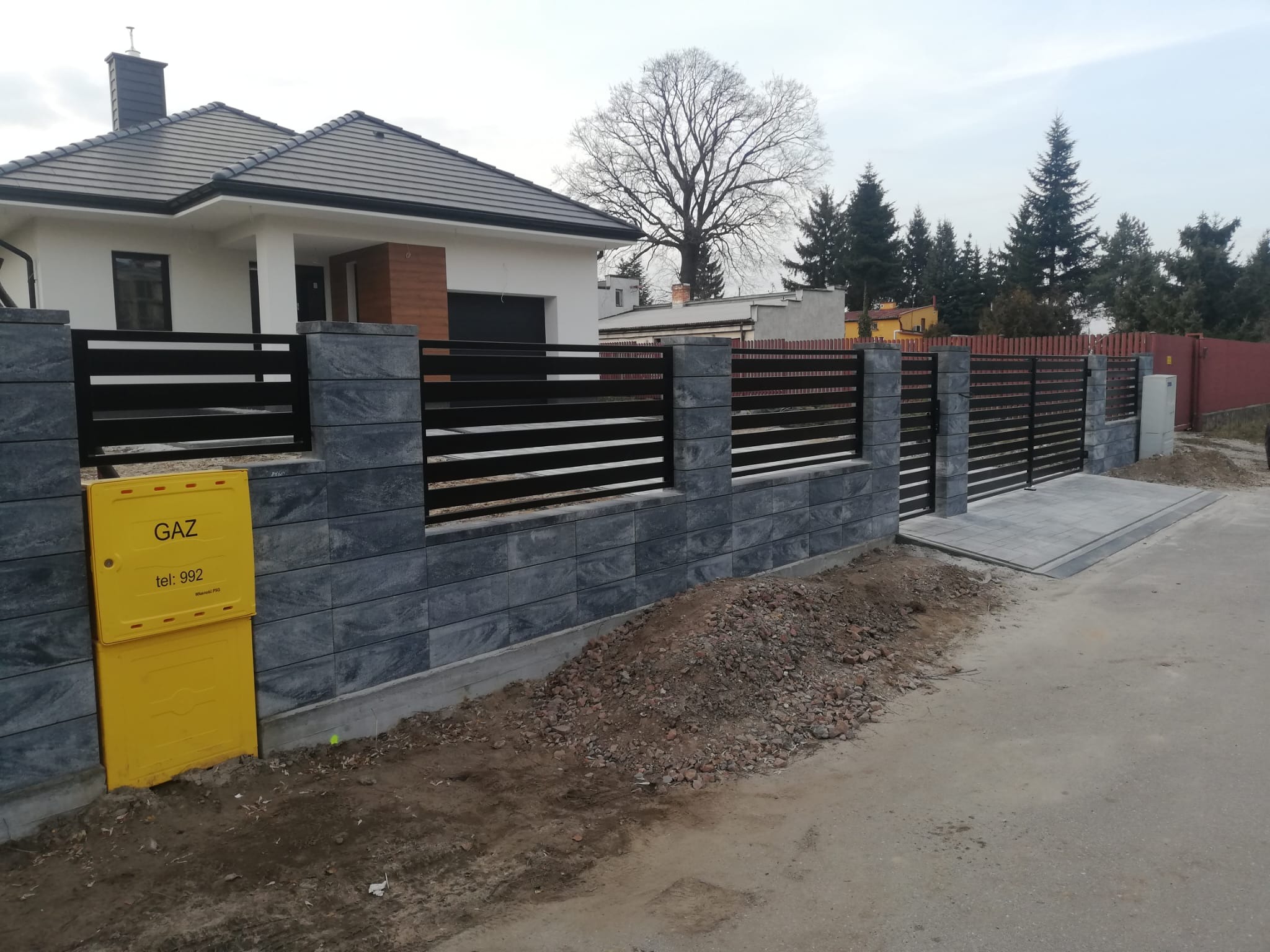 Nowoczesne ogrodzenie posesji z poziomymi, czarnymi przęsłami i szarymi, kamiennymi słupkami, widoczna żółta skrzynka gazowa i fragment nowo wybudowanego domu z szarym dachem.
