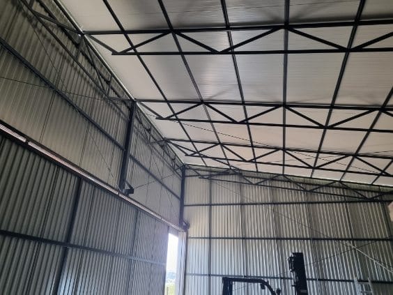 Wnętrze hali z konstrukcją stalową dachu i ścianami z blachy falistej, widoczne okno w oddali.