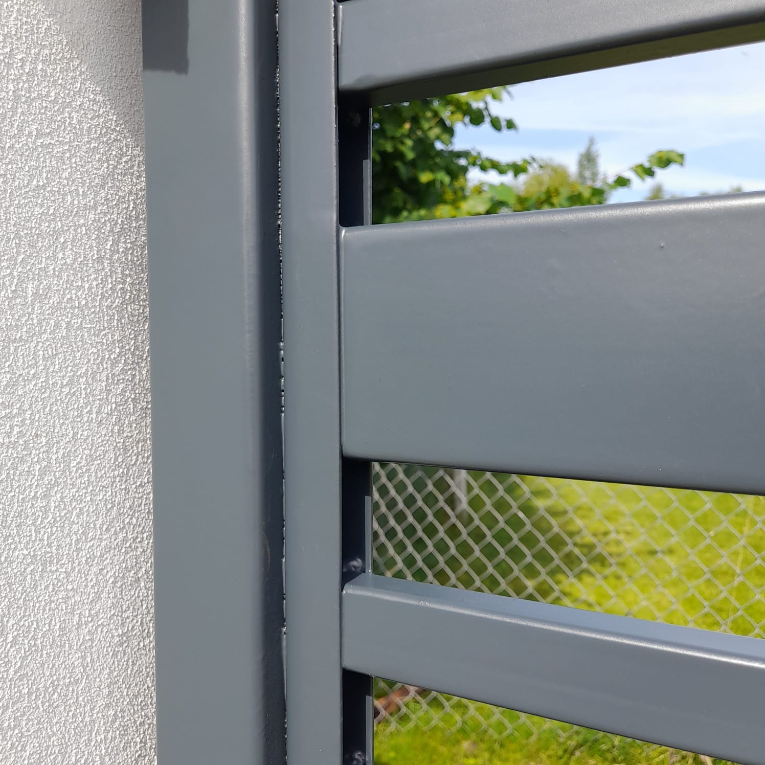 Fragment nowoczesnej, szarej bramy wjazdowej z poziomymi panelami, widok z bliska na tle ściany budynku i zieleni, Radom.