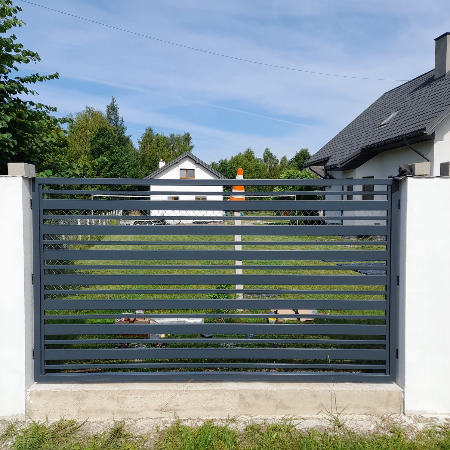 Nowoczesna brama wjazdowa w kolorze grafitowym z poziomymi panelami, widok na dom z ogrodem i pomarańczowy pachołek w tle.