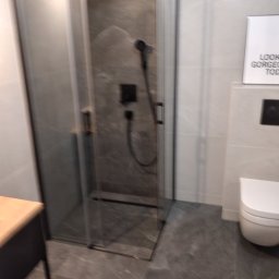 TB Usługi remontowe - Nowoczesna kabina prysznicowa z ciemnoszarą armaturą i płytkami imitującymi kamień w łazience w Zielonej Górze. Na ścianie biała toaleta i plakat z napisem.