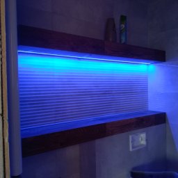 TB Usługi remontowe - Nowoczesna łazienka z podświetlanymi półkami z ciemnego drewna i szarymi płytkami, oświetlona niebieskim światłem LED, z widoczną ścianą w trakcie wykończenia.