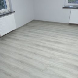 TB Usługi remontowe - Jasne panele podłogowe ułożone w pustym pokoju z białymi ścianami i dwoma oknami, widoczne grzejniki pod oknami.