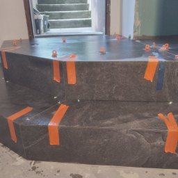 TB Usługi remontowe - Wyłożone ciemnoszarą płytką schody wejściowe do domu, zabezpieczone pomarańczową taśmą i poziomowane plastikowymi krzyżykami, widoczna szpachelka na podłodze.