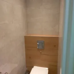 Nowoczesna toaleta podwieszana z beżowymi płytkami imitującymi marmur i drewnianą zabudową stelaża, widoczny chromowany przycisk spłukiwania.