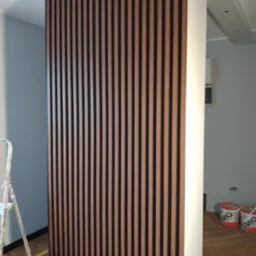 Dekoracyjna ścianka z drewnianych lameli w kolorze orzecha włoskiego z czarnymi wstawkami, zamontowana w rogu pomieszczenia, widoczna drabina malarska i puszki z farbą na podłodze.