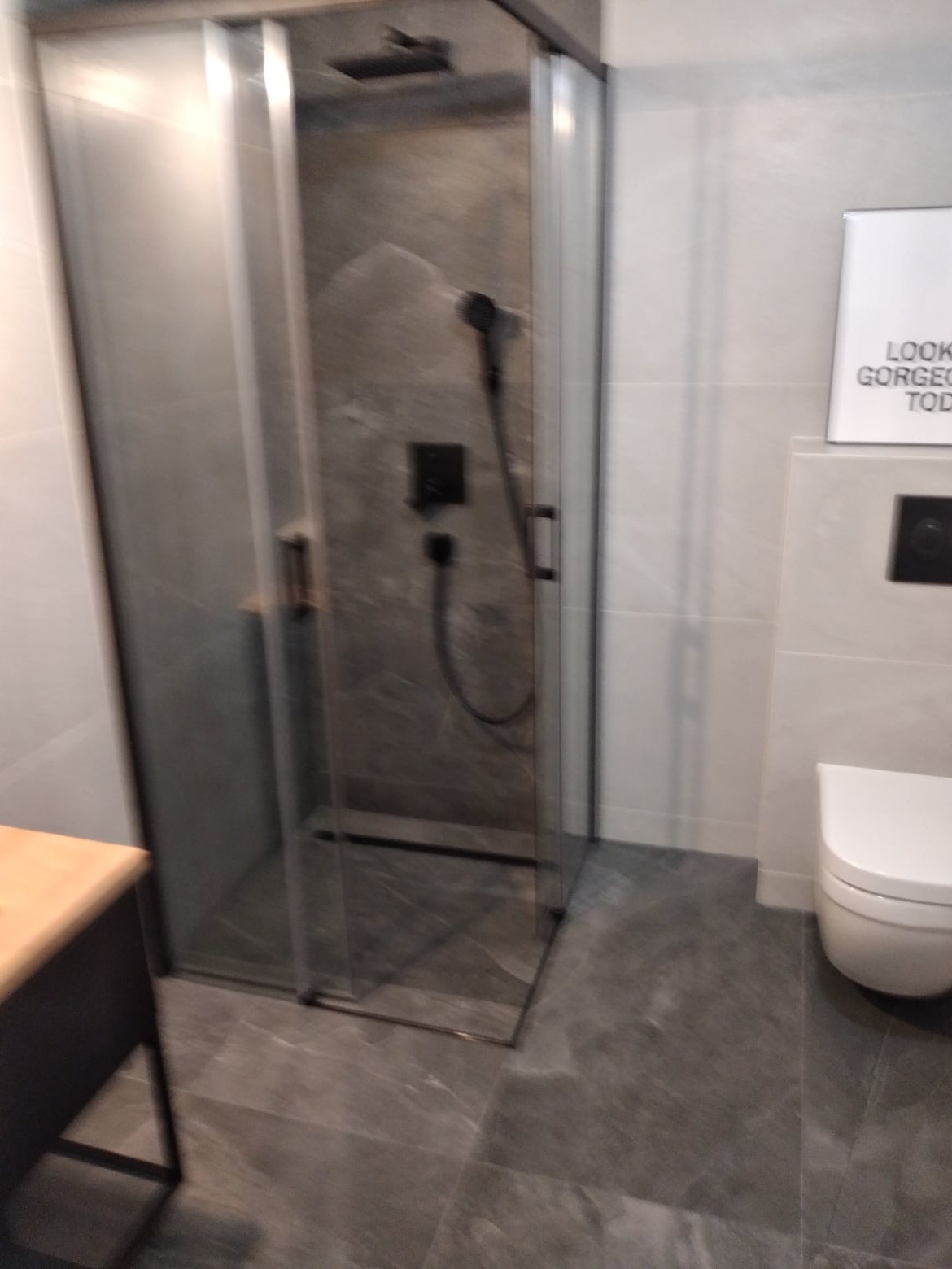 Nowoczesna kabina prysznicowa z ciemnoszarą armaturą i płytkami imitującymi kamień w łazience w Zielonej Górze. Na ścianie biała toaleta i plakat z napisem.