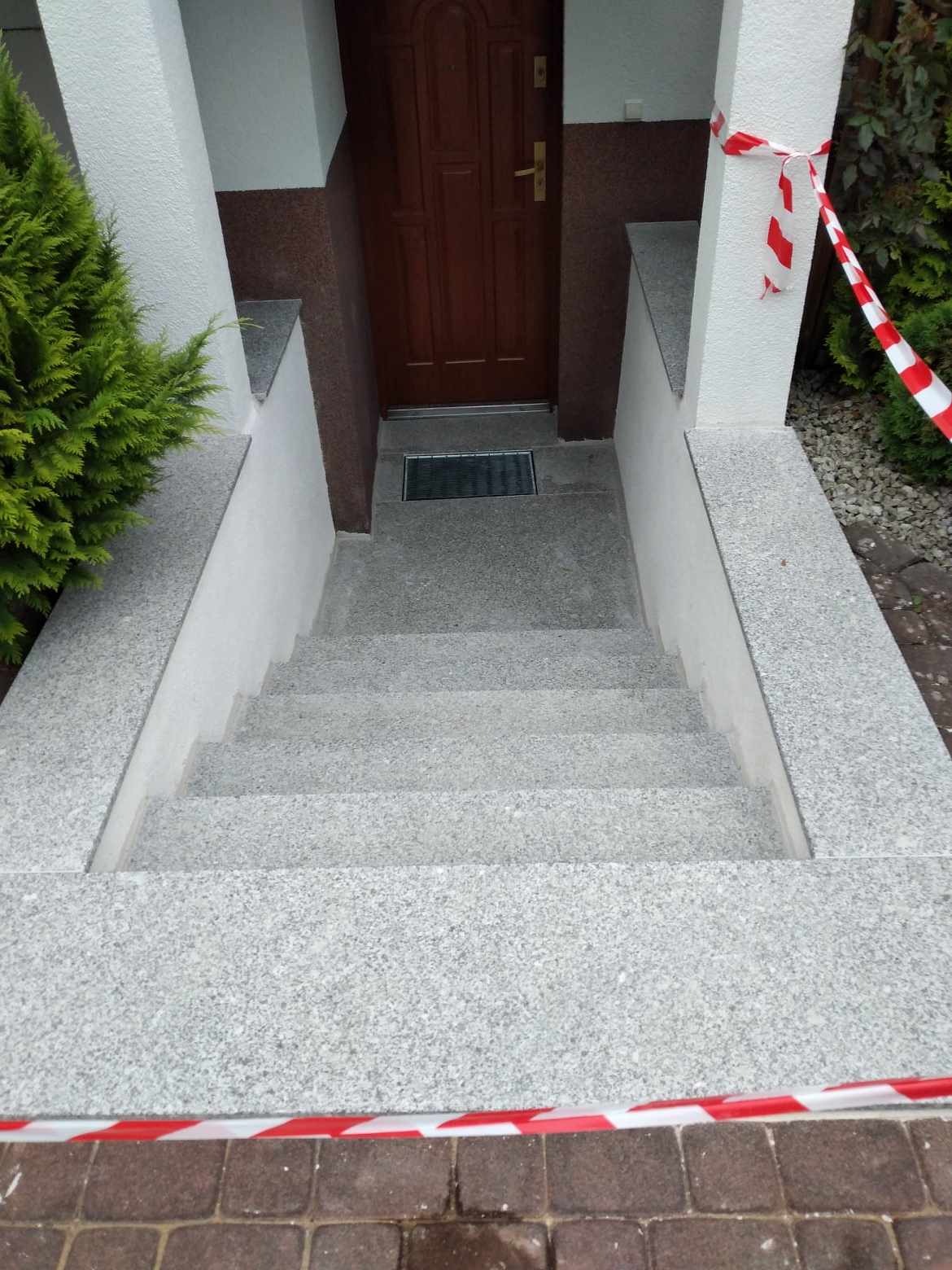 Granitowe schody wejściowe do domu, prowadzące do brązowych drzwi, z widoczną kratką ściekową i taśmą ostrzegawczą.