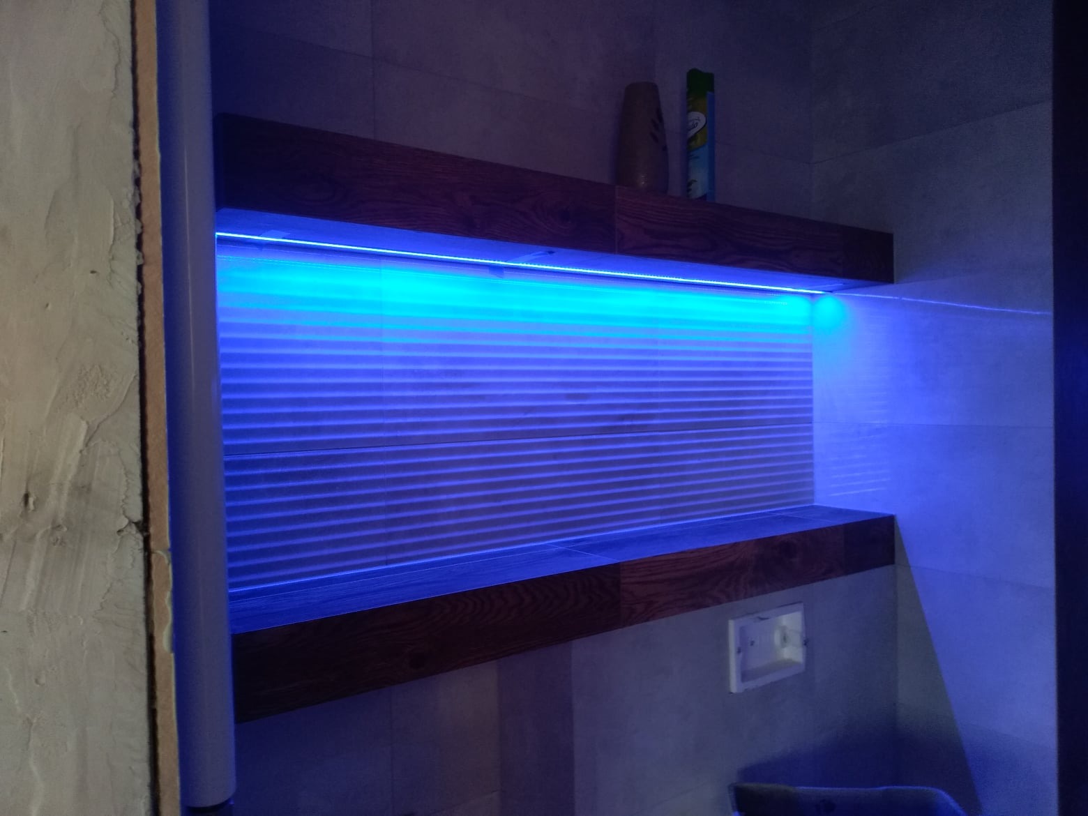 Nowoczesna łazienka z podświetlanymi półkami z ciemnego drewna i szarymi płytkami, oświetlona niebieskim światłem LED, z widoczną ścianą w trakcie wykończenia.