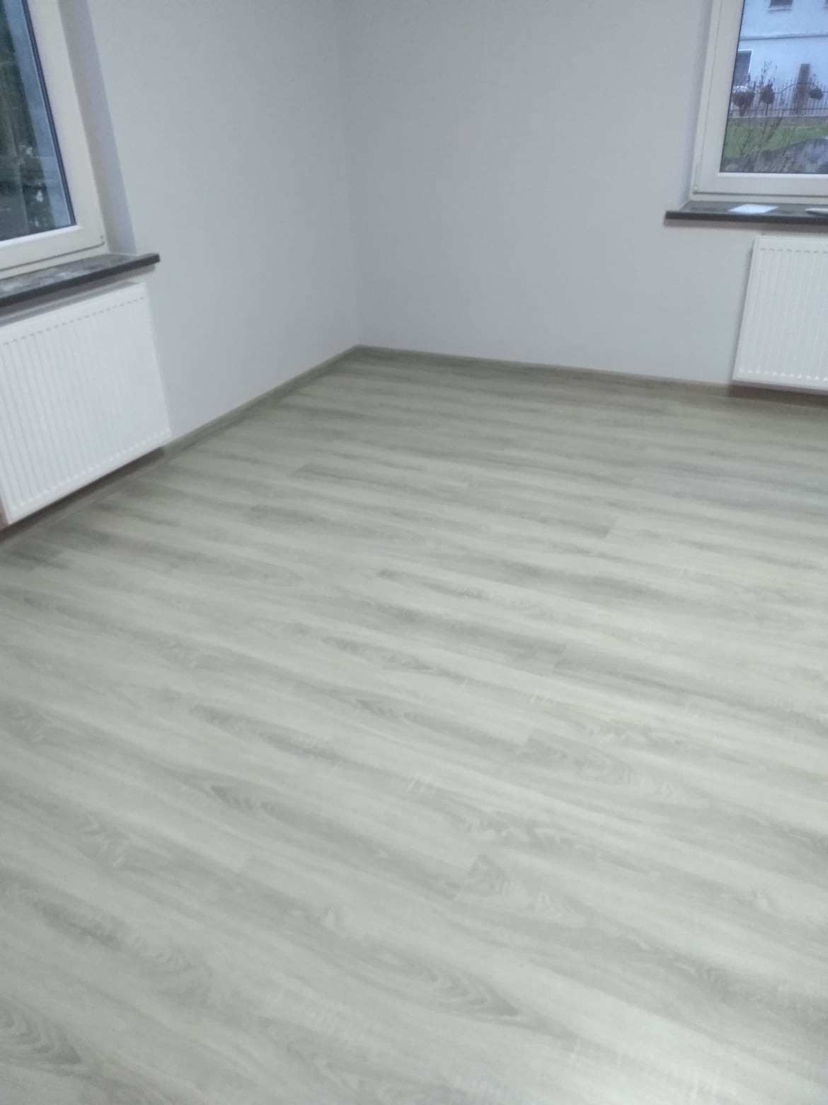 Jasne panele podłogowe ułożone w pustym pokoju z białymi ścianami i dwoma oknami, widoczne grzejniki pod oknami.