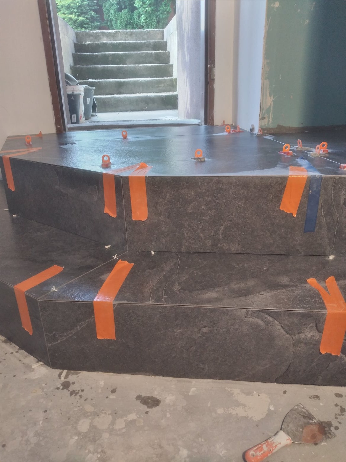 Wyłożone ciemnoszarą płytką schody wejściowe do domu, zabezpieczone pomarańczową taśmą i poziomowane plastikowymi krzyżykami, widoczna szpachelka na podłodze.