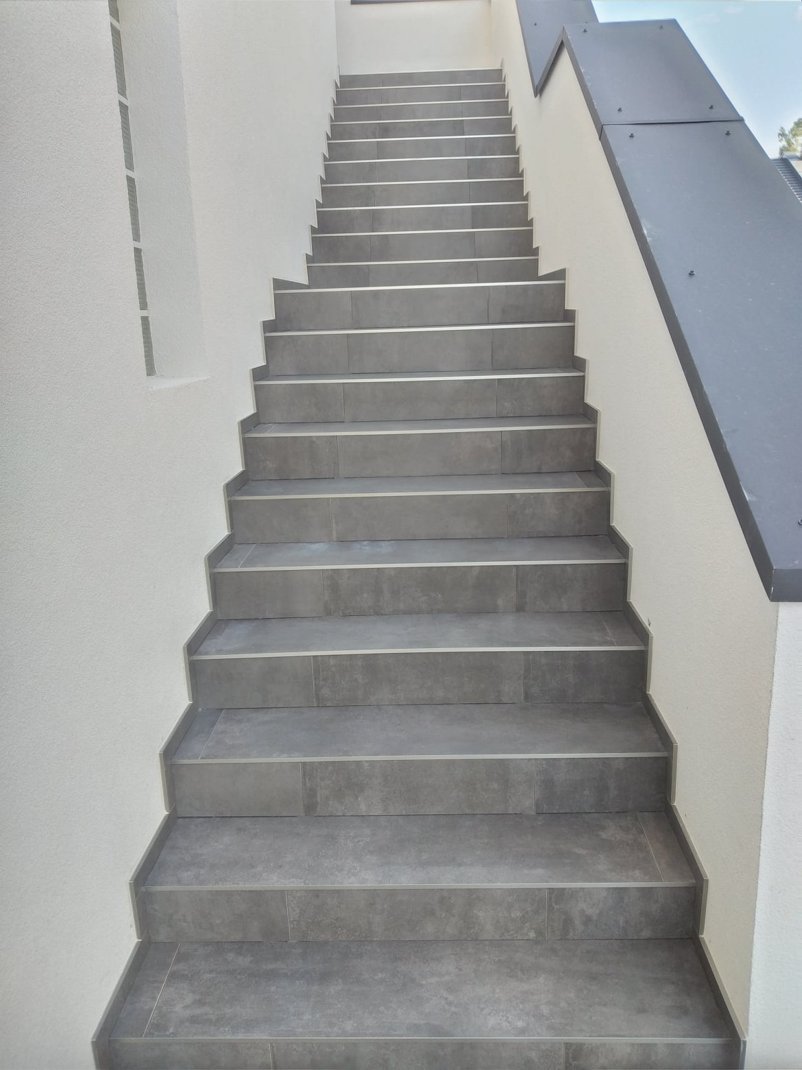 Schody wyłożone szarymi płytkami z delikatnym wzorem, prowadzące w górę, z białymi ścianami po bokach i nowoczesną, ciemnoszarą balustradą po prawej stronie.