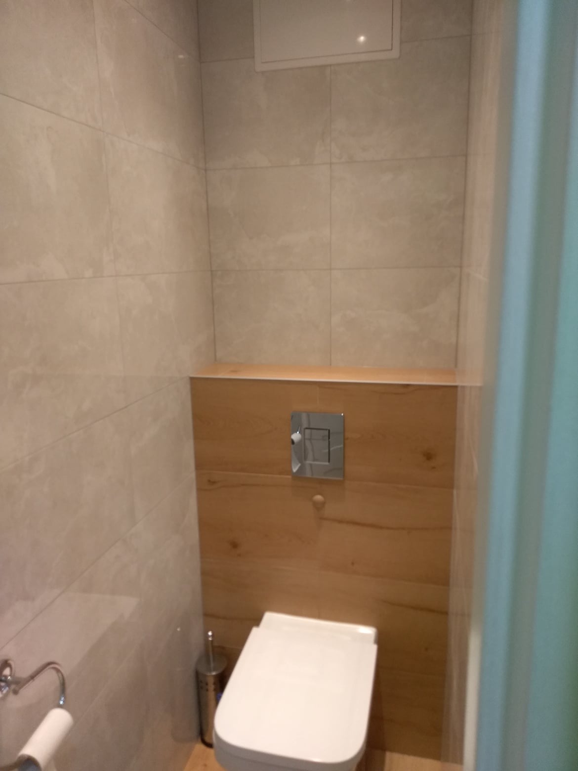 Nowoczesna toaleta podwieszana z beżowymi płytkami imitującymi marmur i drewnianą zabudową stelaża, widoczny chromowany przycisk spłukiwania.