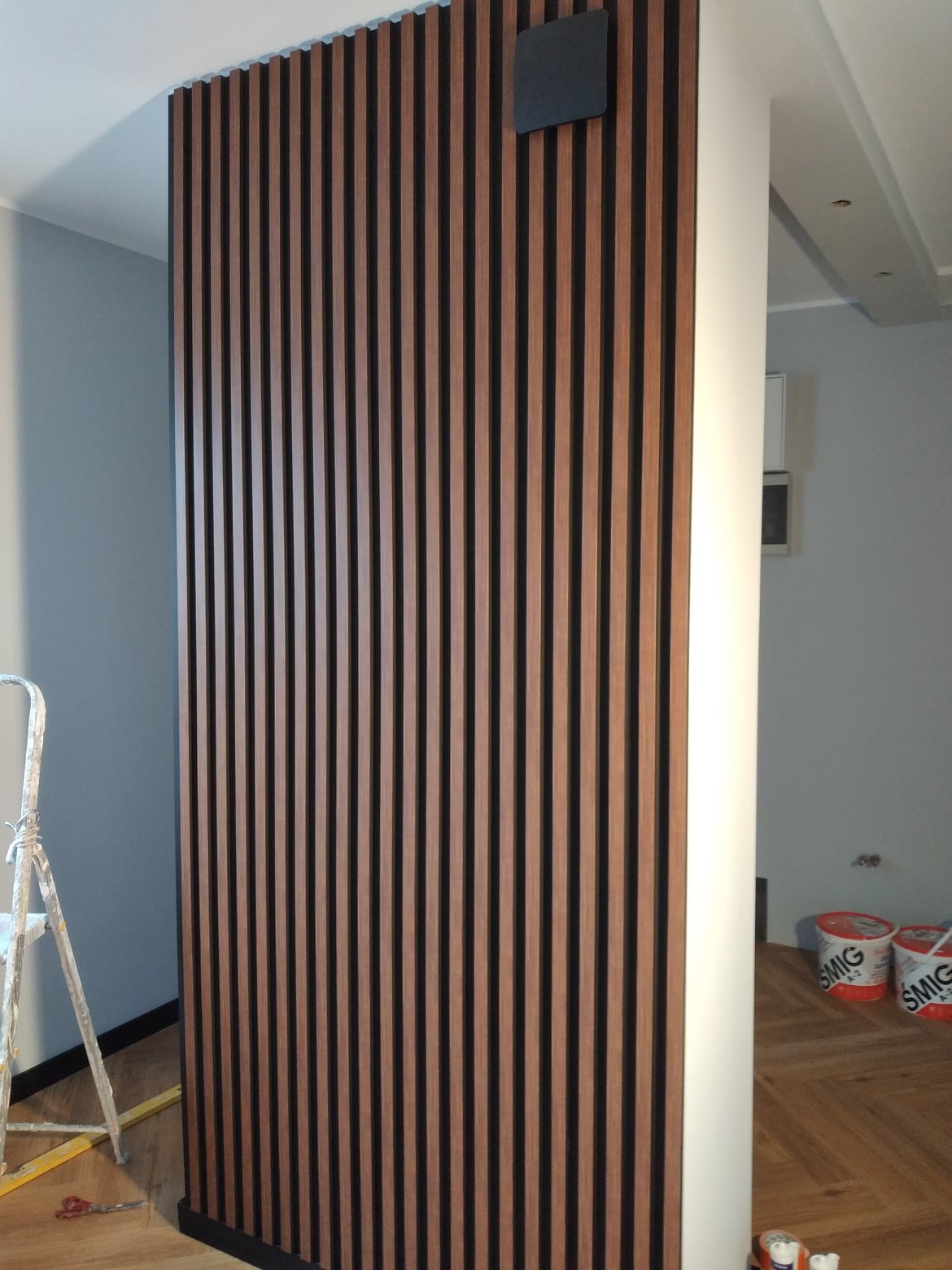 Dekoracyjna ścianka z drewnianych lameli w kolorze orzecha włoskiego z czarnymi wstawkami, zamontowana w rogu pomieszczenia, widoczna drabina malarska i puszki z farbą na podłodze.