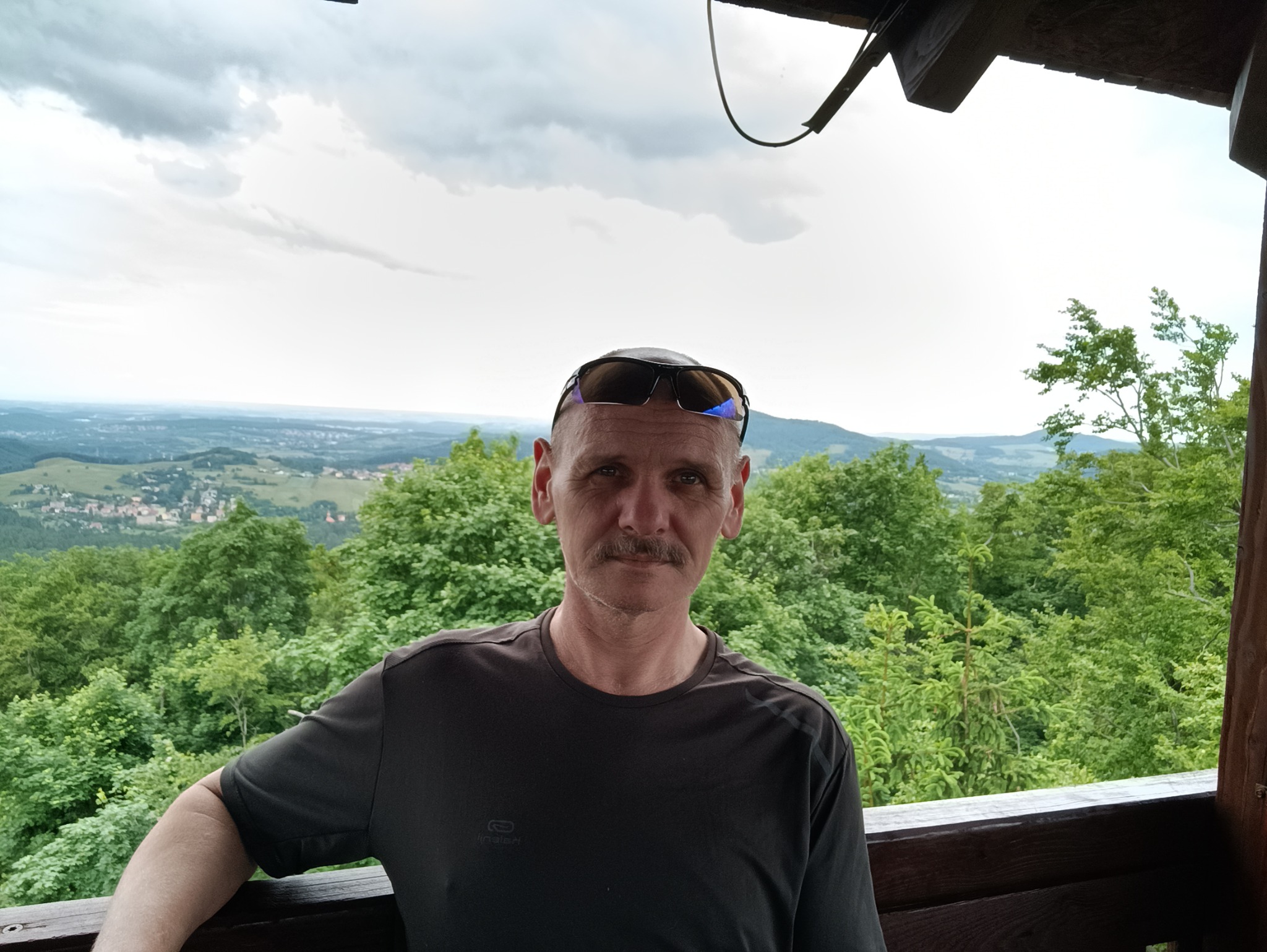 Mężczyzna z wąsami i okularami na głowie, oparty o drewnianą barierkę z widokiem na zalesione wzgórza i miasto w oddali, pochmurne niebo.