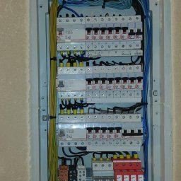 Usługi elektryczne i ogólnobudowlane Rafał Turski - Rozdzielnica elektryczna z zainstalowanymi bezpiecznikami i przewodami w kolorach niebieskim, żółtym i zielonym, zamontowana we wnęce w surowej ścianie.