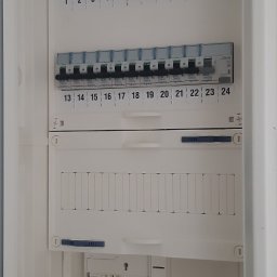 Usługi elektryczne i ogólnobudowlane Rafał Turski - Rozdzielnica elektryczna w białej obudowie, widoczne bezpieczniki z numeracją, cztery czerwone moduły ochronne. Dolna część obudowy otwarta, ukazująca okablowanie.