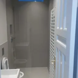 Nowoczesna łazienka z szarą glazurą, deszczownicą, wiszącą toaletą i umywalką nablatową, widok na białe drzwi z ozdobną kratką wentylacyjną.