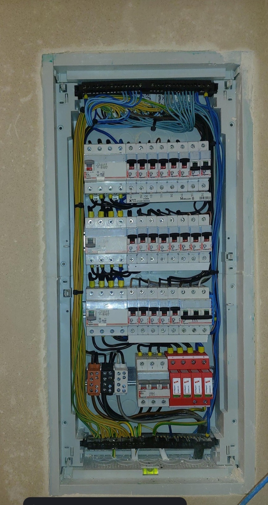 Rozdzielnica elektryczna z zainstalowanymi bezpiecznikami i przewodami w kolorach niebieskim, żółtym i zielonym, zamontowana we wnęce w surowej ścianie.