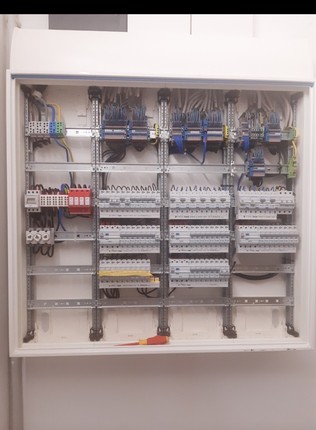 Otwarta skrzynka rozdzielcza z zamontowanymi bezpiecznikami, przewodami i szynami DIN, widoczny pomarańczowo-żółty śrubokręt na dnie obudowy.
