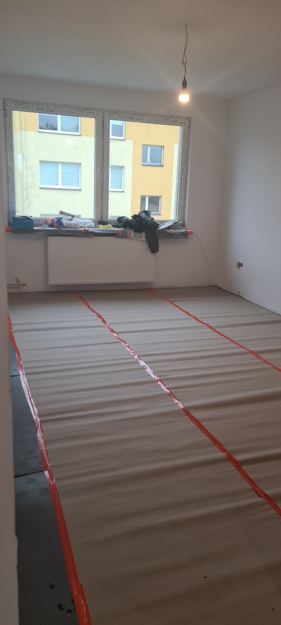 Pokój w trakcie przygotowania do remontu: podłoga zabezpieczona kartonem i taśmą, widoczne narzędzia i materiały na parapecie, surowa żarówka zwisająca z sufitu.