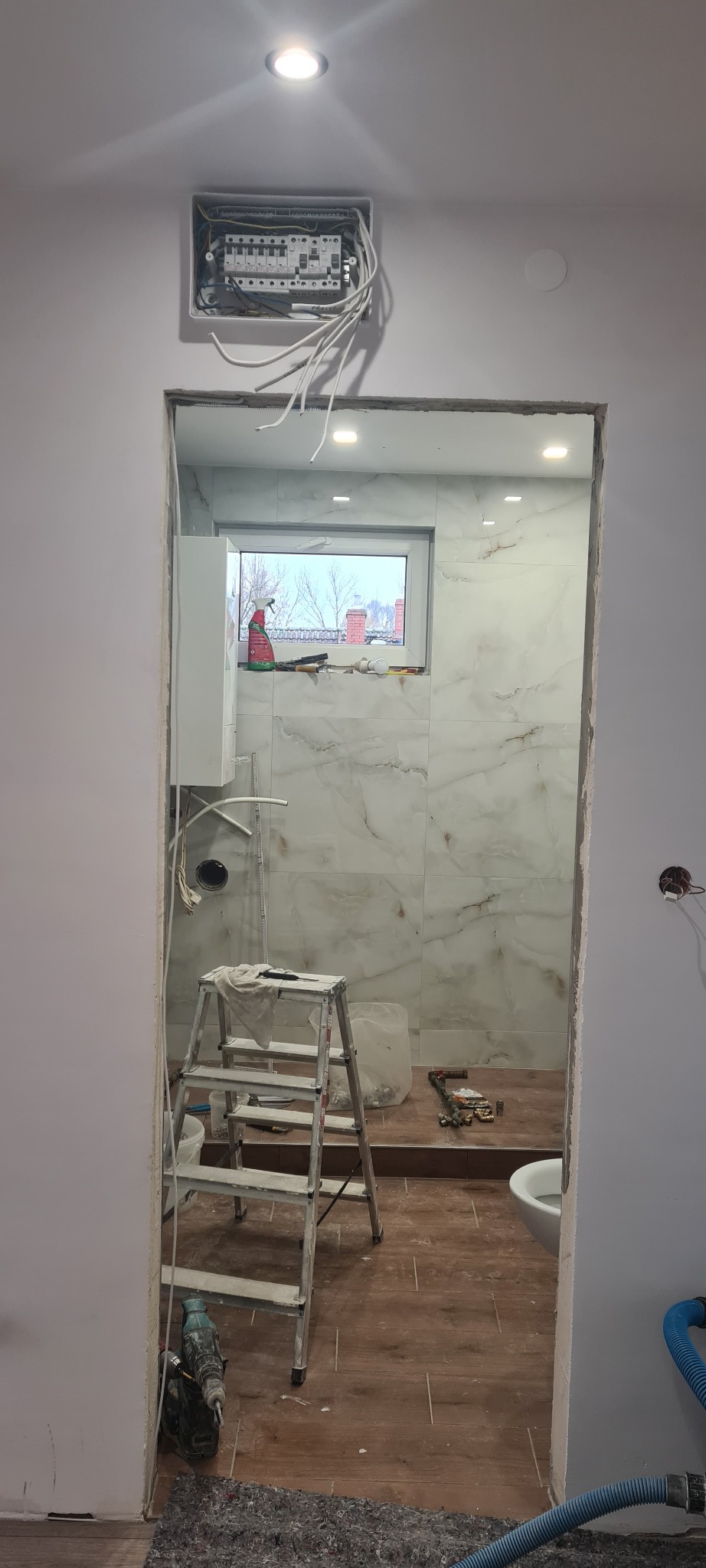 Remontowana łazienka z marmurowymi płytkami, widoczna drabina, narzędzia, niedokończona instalacja elektryczna i okno z widokiem na zimowy krajobraz.