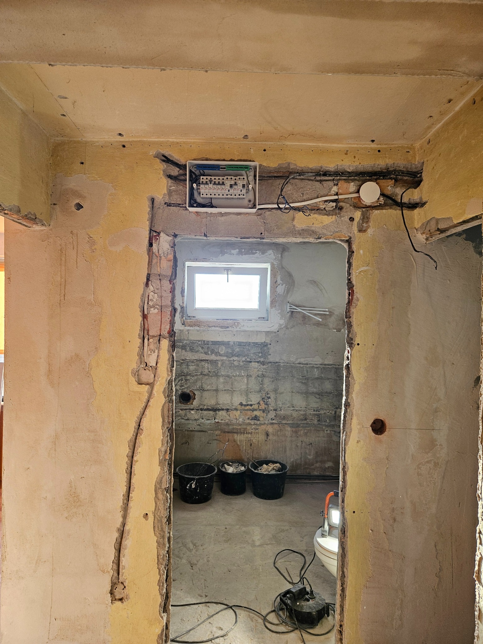 Wnętrze w trakcie remontu: widoczne ściany ze śladami skuwania, puszka z bezpiecznikami nad otworem drzwiowym, kable, wiadra z zaprawą i lampa budowlana w tle.