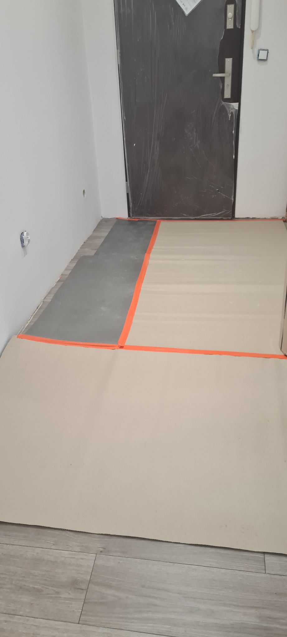 Przygotowanie podłoża pod nową podłogę: wyrównana wylewka częściowo zakryta folią ochronną, zabezpieczona pomarańczową taśmą malarską przy drzwiach wejściowych i ścianie.