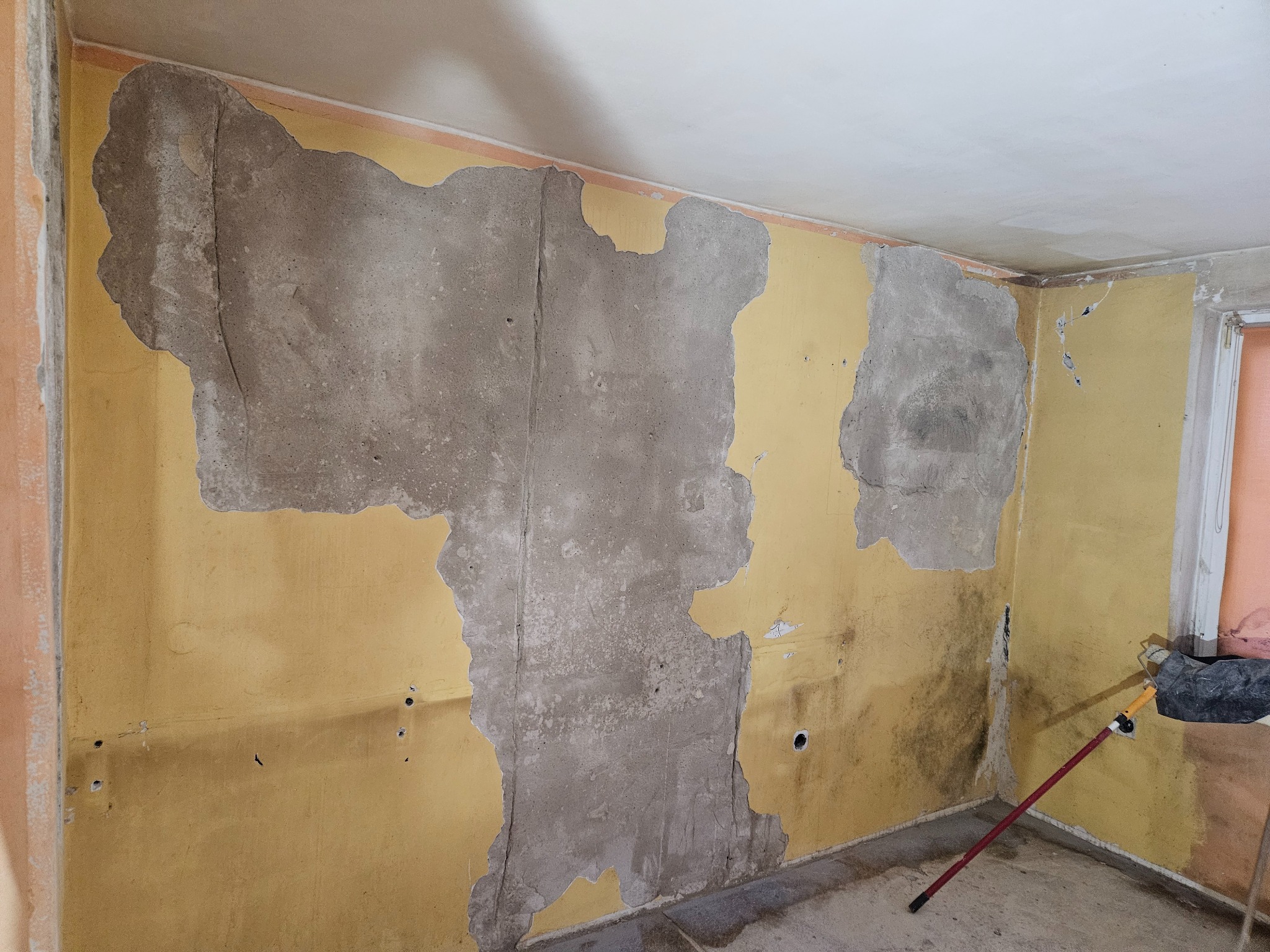 Żółta ściana w pokoju z fragmentami starego tynku, odkrywająca szary beton pod spodem, widoczne ślady po wierceniach, w rogu pomieszczenia wiadro i wałek malarski.