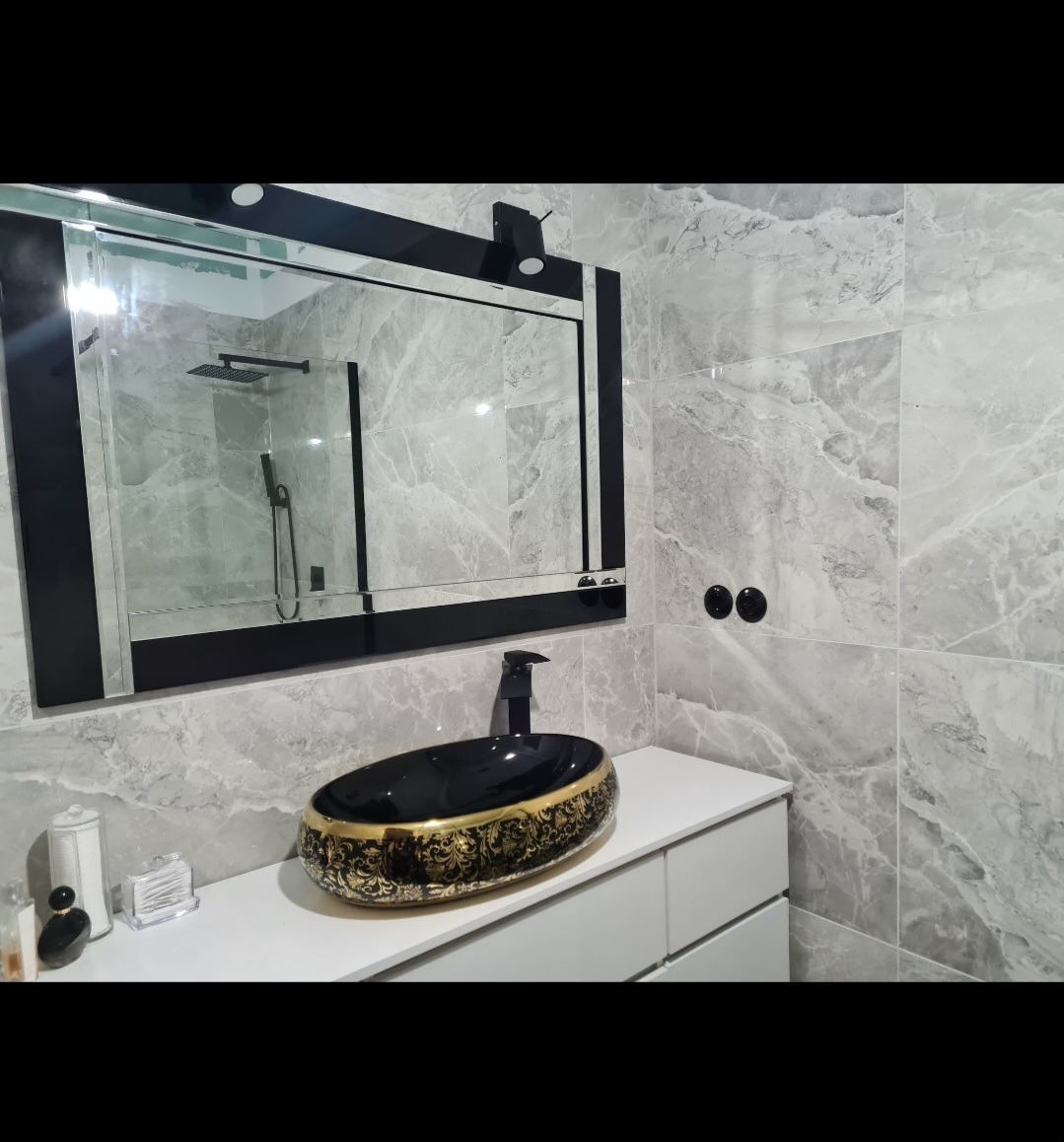 Wykończona łazienka z umywalką nablatową w kolorze czarnym ze złotymi zdobieniami, duże lustro w czarnej ramie, szare płytki imitujące marmur na ścianach.