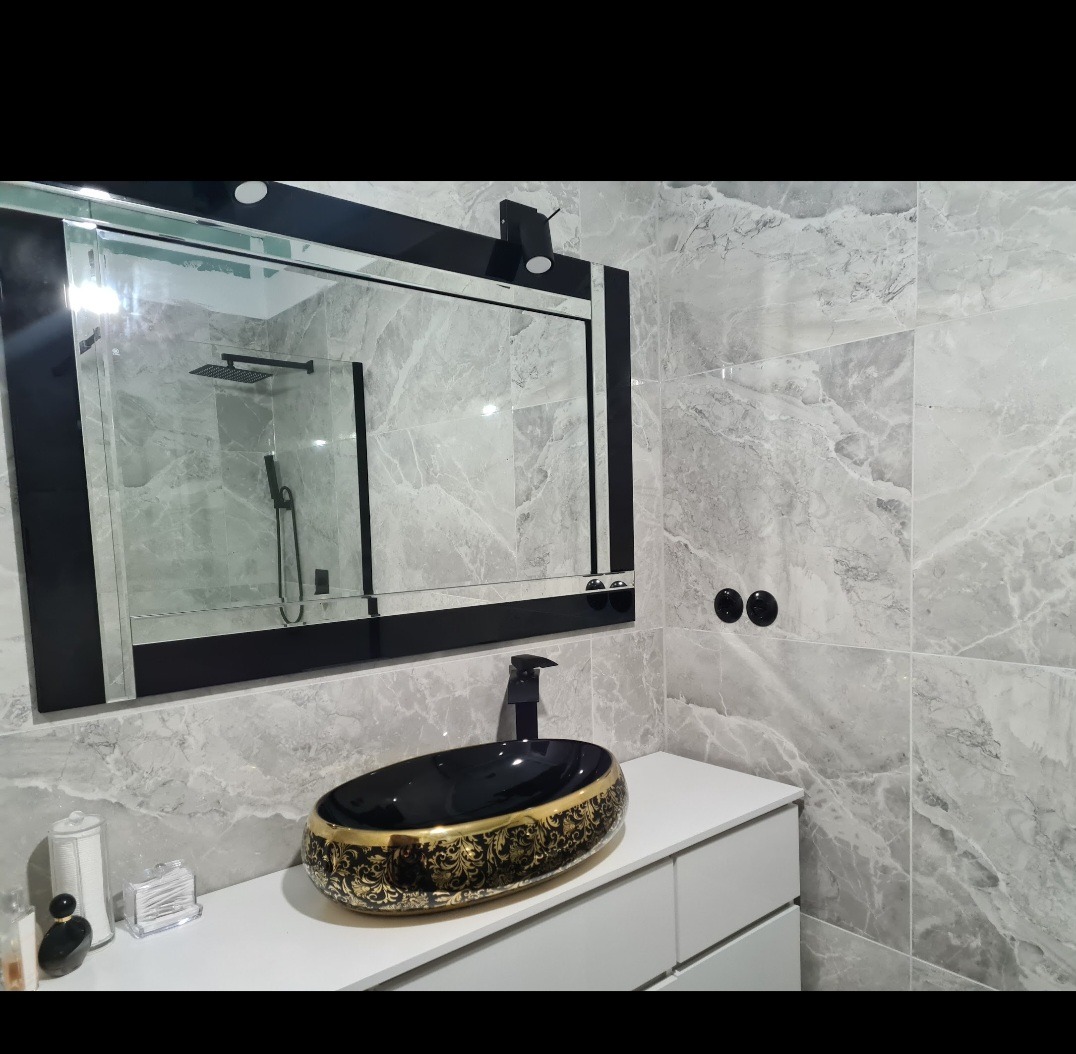 Nowoczesna łazienka z umywalką nablatową w kolorze czarnym ze złotymi zdobieniami, czarną armaturą i dużym lustrem w czarnej ramie, na tle płytek imitujących marmur.