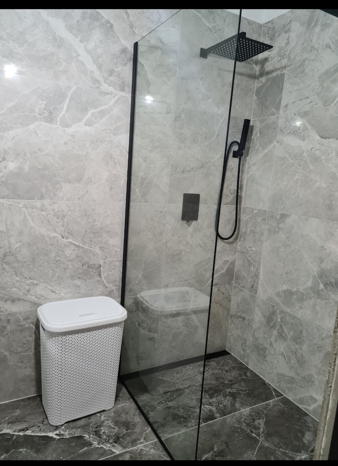 Nowoczesna kabina prysznicowa z czarną armaturą i szarymi płytkami imitującymi marmur. W rogu stoi biały kosz na pranie z tworzywa.