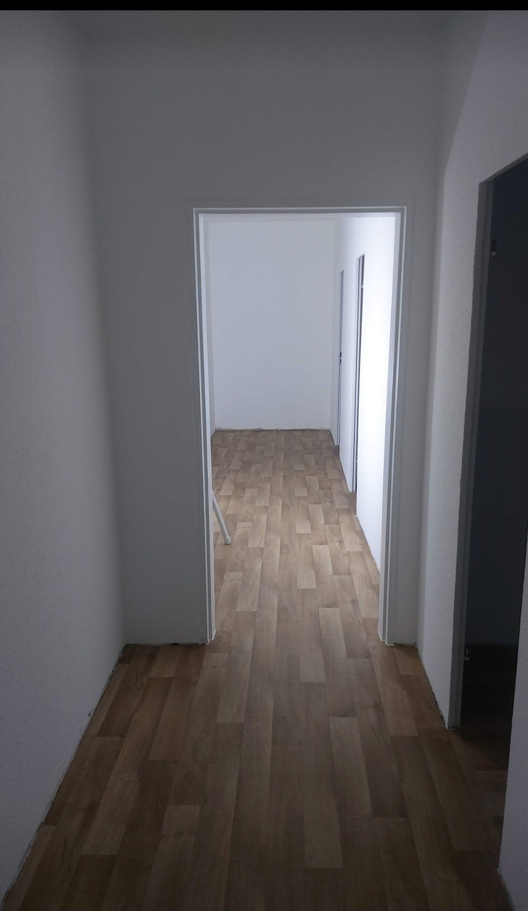 Korytarz z nowo położoną podłogą z paneli imitujących drewno, widok przez otwarte drzwi do kolejnego pomieszczenia, ściany pomalowane na biało.