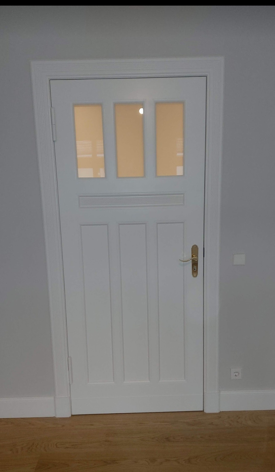 Białe, przeszklone drzwi wewnętrzne z ozdobną listwą i złotą klamką, zamontowane w szarej ścianie z widocznym gniazdkiem i listwą przypodłogową.