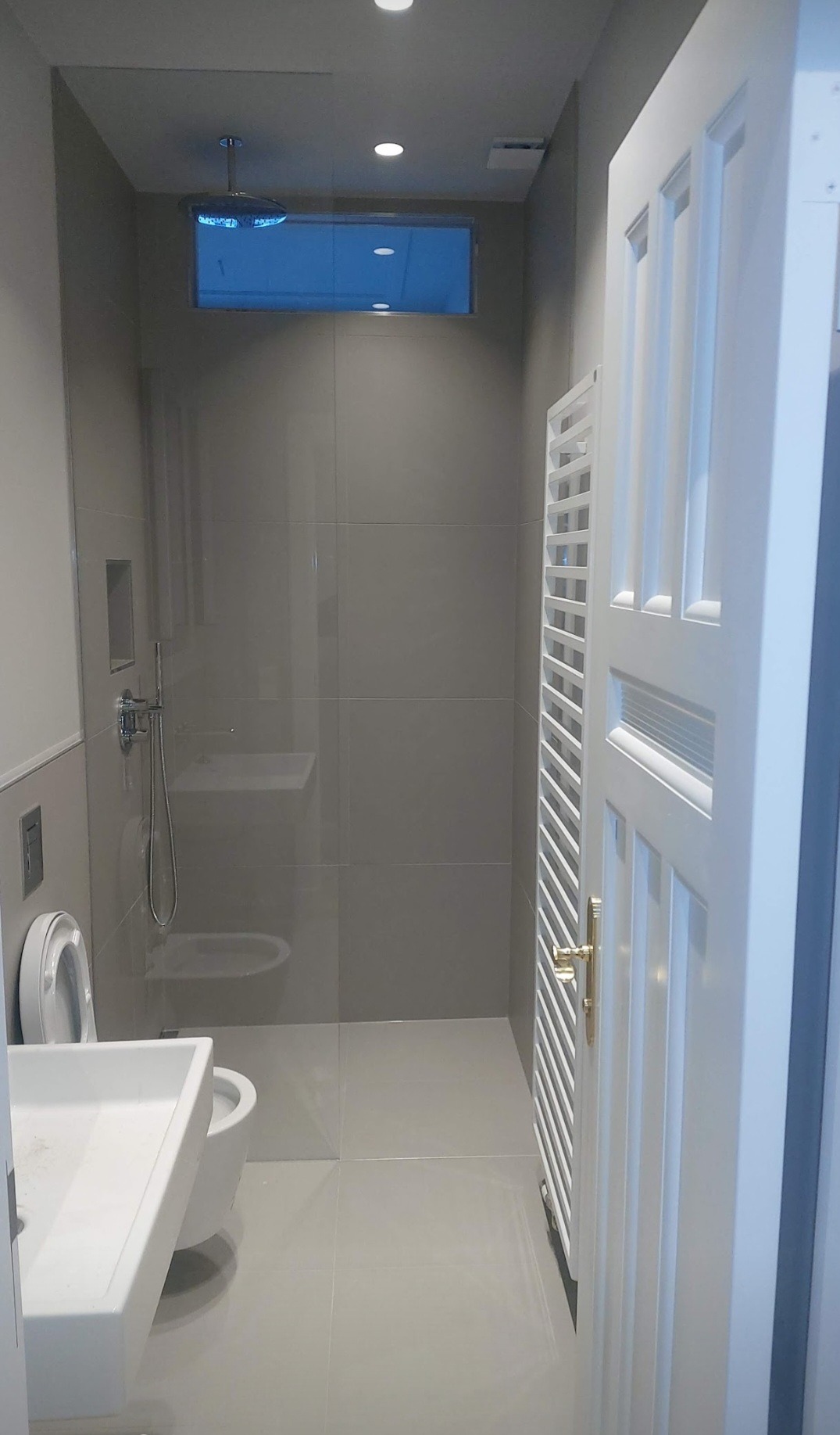 Nowoczesna łazienka z szarą glazurą, deszczownicą, wiszącą toaletą i umywalką nablatową, widok na białe drzwi z ozdobną kratką wentylacyjną.