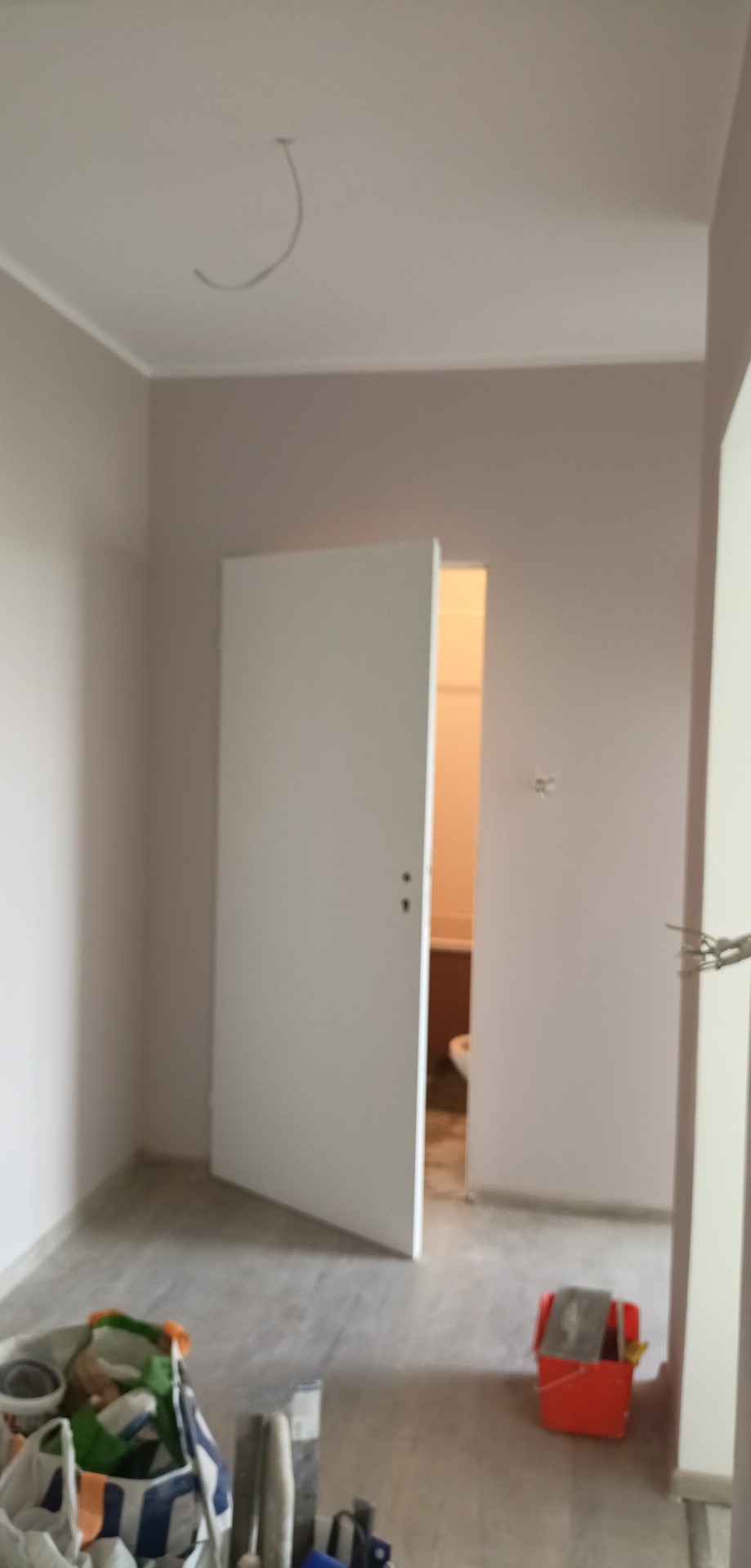 Pokój w trakcie malowania, z widocznymi narzędziami malarskimi, otwartymi drzwiami do łazienki i świeżo pomalowanymi ścianami w odcieniach beżu.