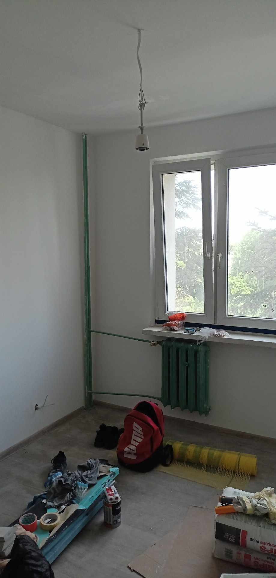 Pokój w trakcie przygotowań do prac wykończeniowych: widoczne narzędzia, materiały budowlane, zabezpieczenia podłogi i grzejnik pomalowany na zielono.