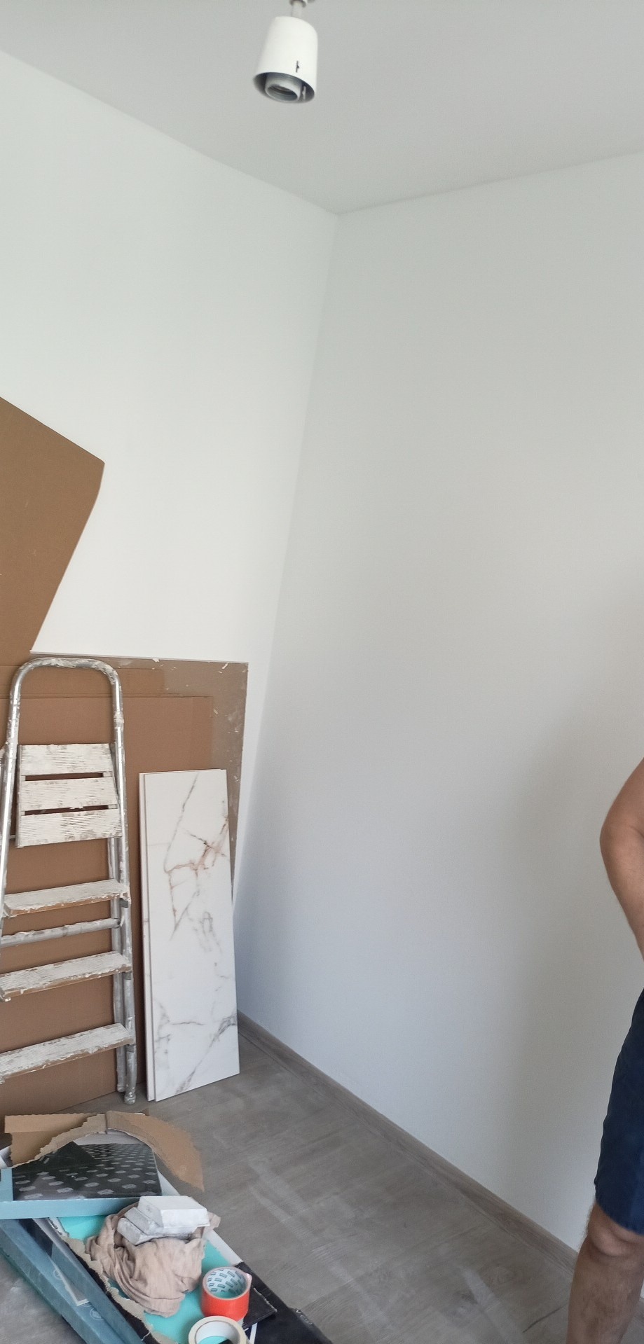 Wnętrze pokoju w trakcie remontu: białe ściany, drabina malarska, płyta imitująca marmur oparta o ścianę, kartony, narzędzia i materiały budowlane na podłodze, widoczny fragment osoby w szortach.