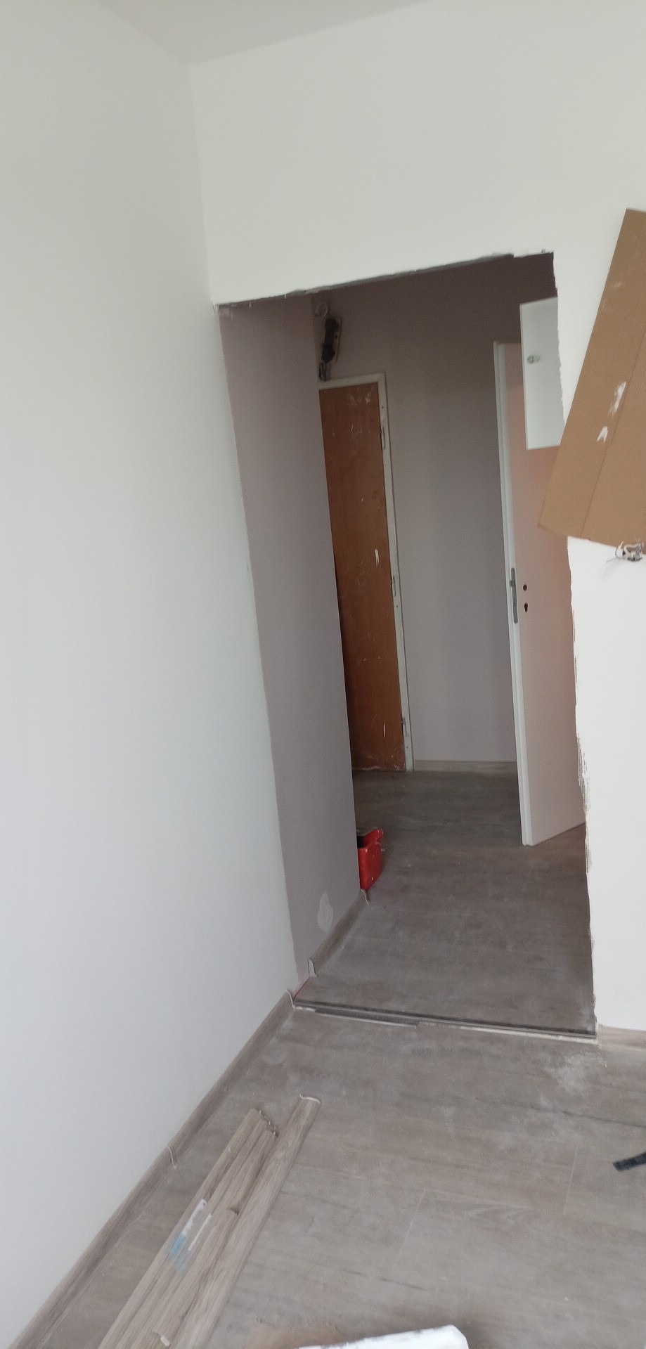 Wnętrze w trakcie prac: świeżo pomalowane ściany na biało, ułożone panele podłogowe w odcieniach szarości, widoczne listwy przypodłogowe i czerwona skrzynka narzędziowa w przejściu do kolejnego...