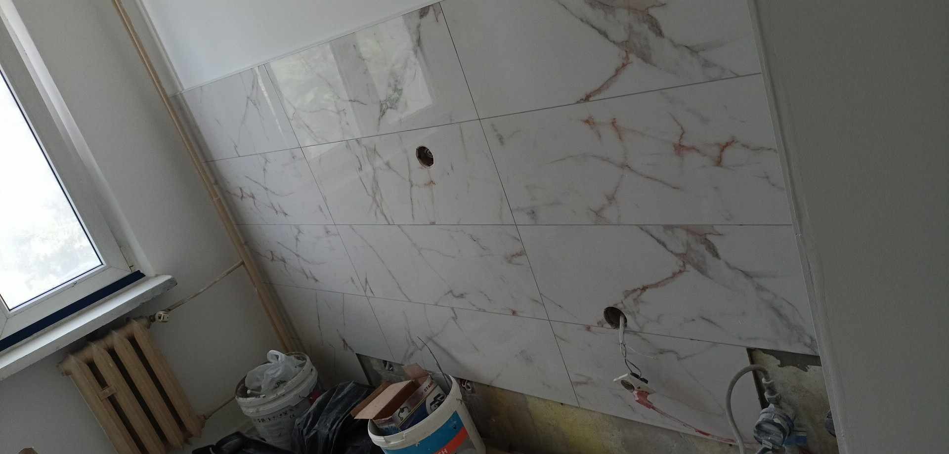 Ściana wyłożona białymi płytkami imitującymi marmur z wywierconymi otworami na gniazdka elektryczne, widoczne wiadra z farbą i narzędzia malarskie w pomieszczeniu w trakcie prac wykończeniowych.