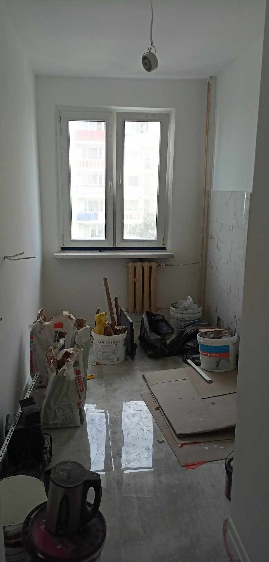 Pokój w trakcie prac przygotowawczych do malowania: worki z gipsem, wiadra z farbą, kartony na podłodze, grzejnik pod oknem, surowe ściany i wystający kabel od lampy.