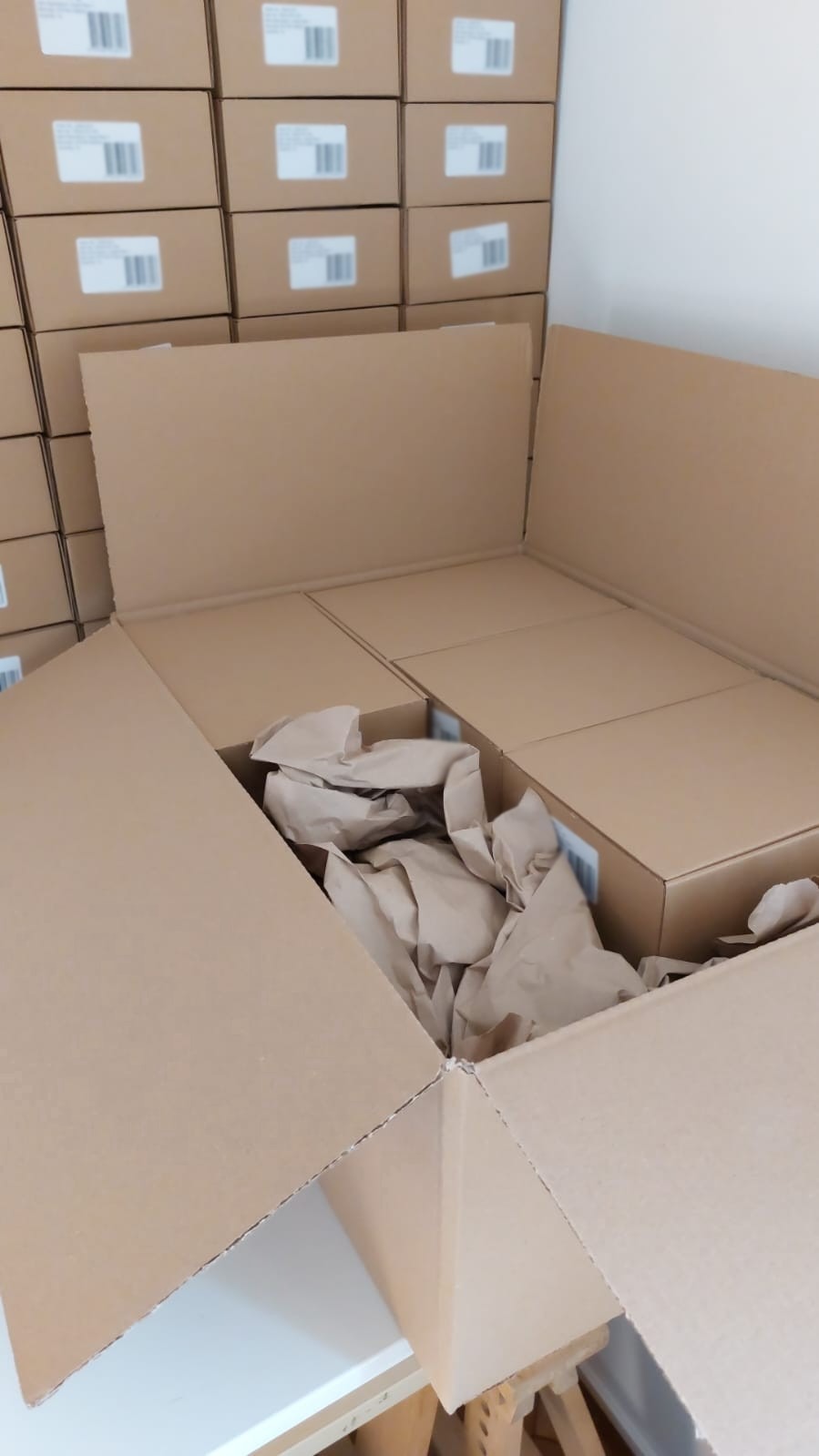 Kartonowe pudła wypełnione papierowym wypełniaczem, ułożone w otwartym kartonie na blacie, w tle stosy identycznych zapakowanych pudeł.