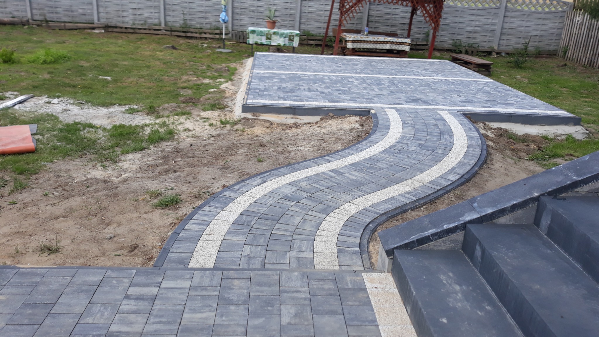 Świeżo ułożona, kręta ścieżka z szarej kostki brukowej z jasnymi paskami, prowadząca do tarasu z kostki, widok z góry schodów, w tle trawnik i ogrodzenie.