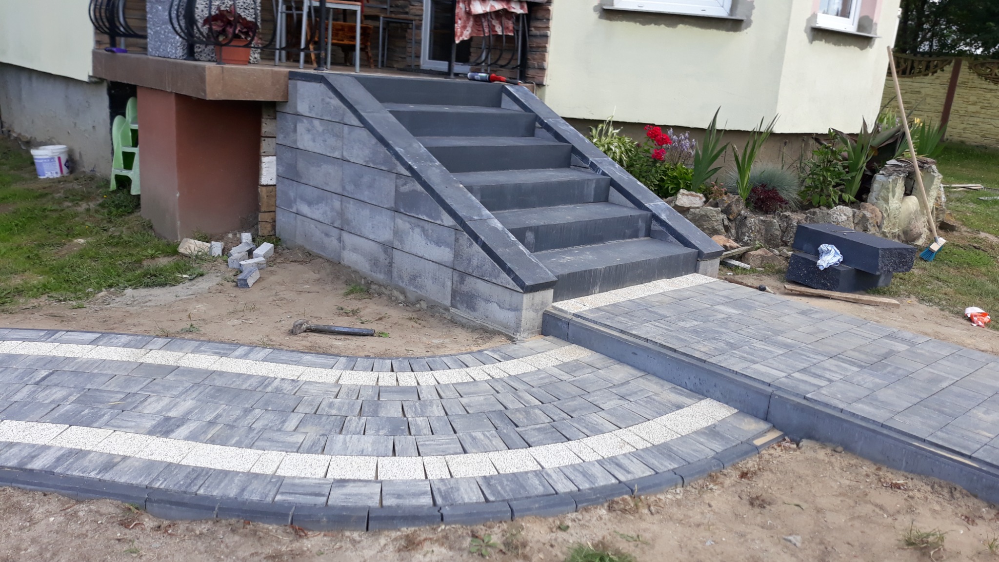 Schody wejściowe do domu wyłożone szarą kostką brukową, z chodnikiem z kostki w dwóch odcieniach szarości, ułożonym w łuk i z obrzeżami z ciemnej kostki