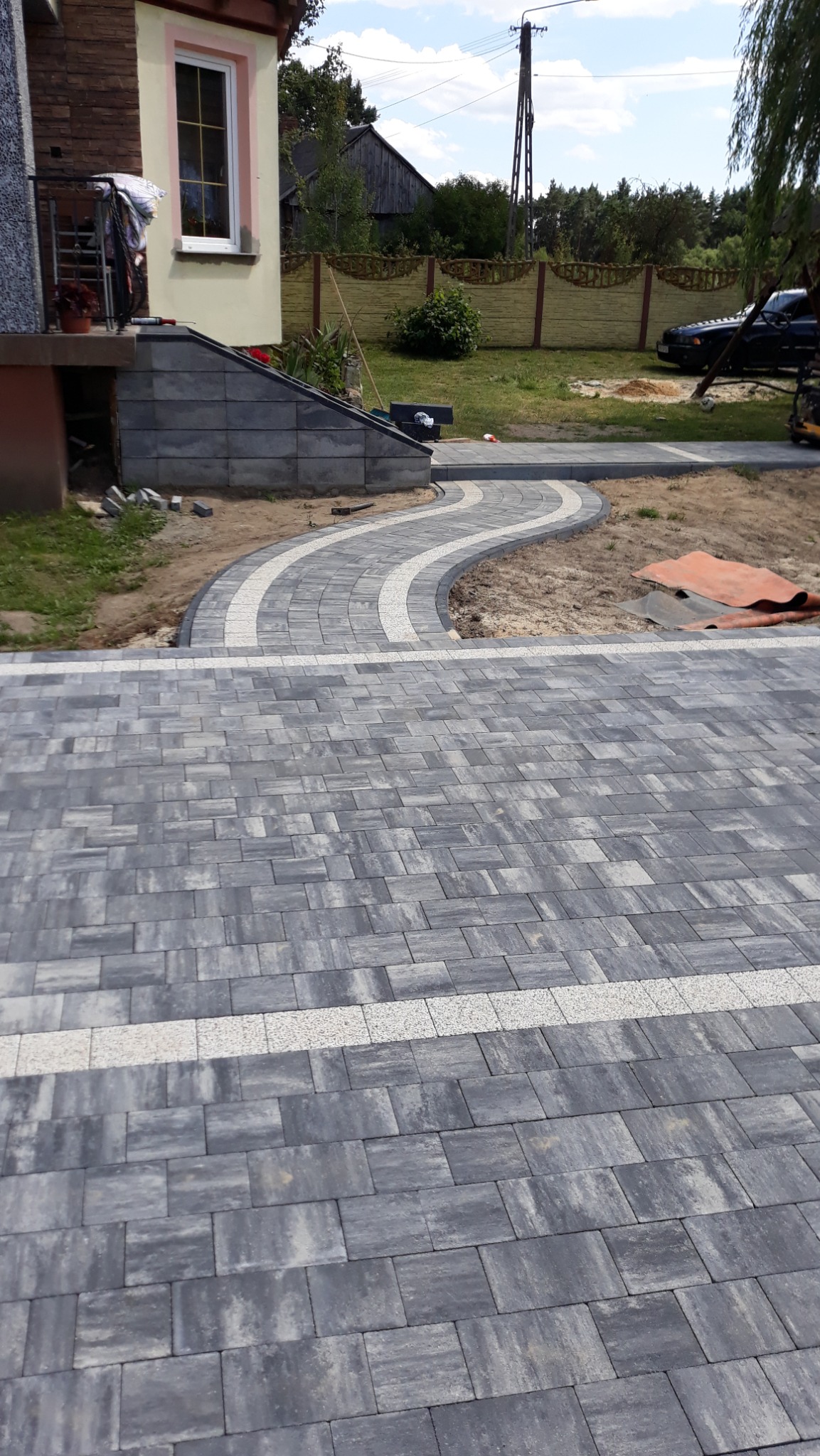 Ułożona szara kostka brukowa z białymi liniami oddzielającymi, tworząca ścieżkę w ogrodzie, widok z góry na posesję z domem i ogrodzeniem w tle.