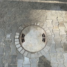 Mikolo Logistic - Szara, betonowa studzienka kanalizacyjna z czterema otworami, otoczona kostką brukową ułożoną w okrąg, z widocznymi śladami piasku i ziemi na powierzchni.