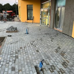 Mikolo Logistic - Szara kostka brukowa ułożona przed budynkiem z żółtą elewacją, widoczne otwarte drzwi i okna w folii ochronnej, w tle robotnik z taczką i wykopane miejsce na kable.