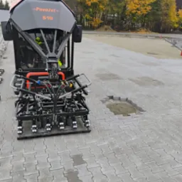 Maszyna PaveJet S19 układająca szarą kostkę brukową typu 'I'. Widoczny fragment terenu przygotowany pod kostkę oraz operator maszyny w odblaskowej kamizelce. W tle drzewa z jesiennymi liśćmi.