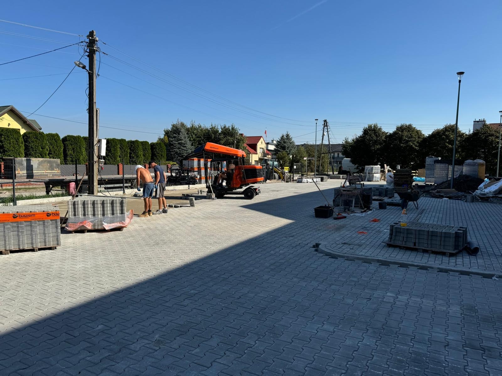 Dwóch mężczyzn w letnich ubraniach układa szarą kostkę brukową na placu, widoczny podnośnik do kostki brukowej, palety z kostką, ułożona nawierzchnia i budynki w tle w słoneczny dzień.