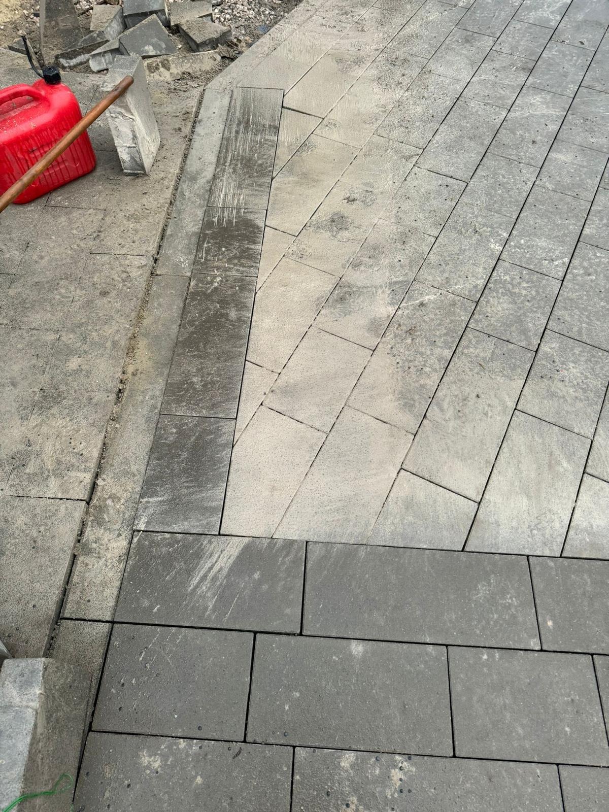Fragment nowo układanej nawierzchni z kostki granitowej w dwóch odcieniach szarości, widoczny kanister i łopata, etap prac w toku.